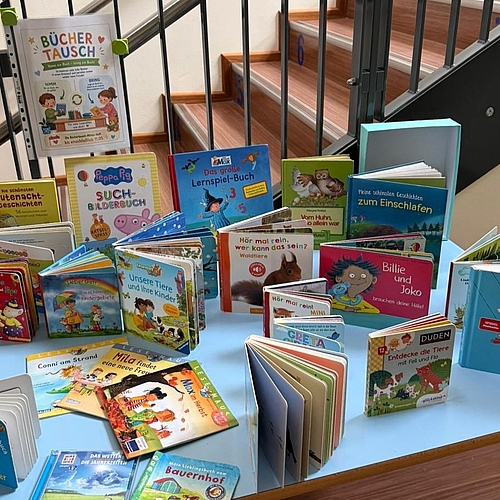 Ein Tisch, der mit vielen Kinderbüchern in verschiedenen Farben und Größen bedeckt ist. Im oberen Bereich ist ein kleines weißes Schild mit der Aufschrift „Büchertausch“ zu sehen. Im Hintergrund ist eine Treppe erkennbar.