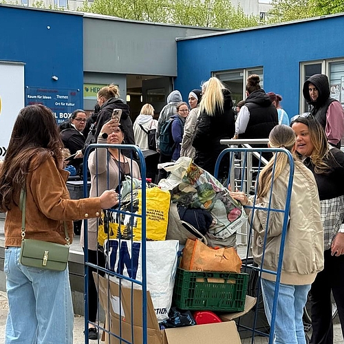 Mehrere Personen stehen im Freien um einen Rollwagen herum, der mit großen Säcken und Tüten voller Bücher und Kleiderspenden beladen ist, bereit für den Transport zur Aktion „Herz im Kiez“. Im Hintergrund ist ein Gebäude zu sehen.